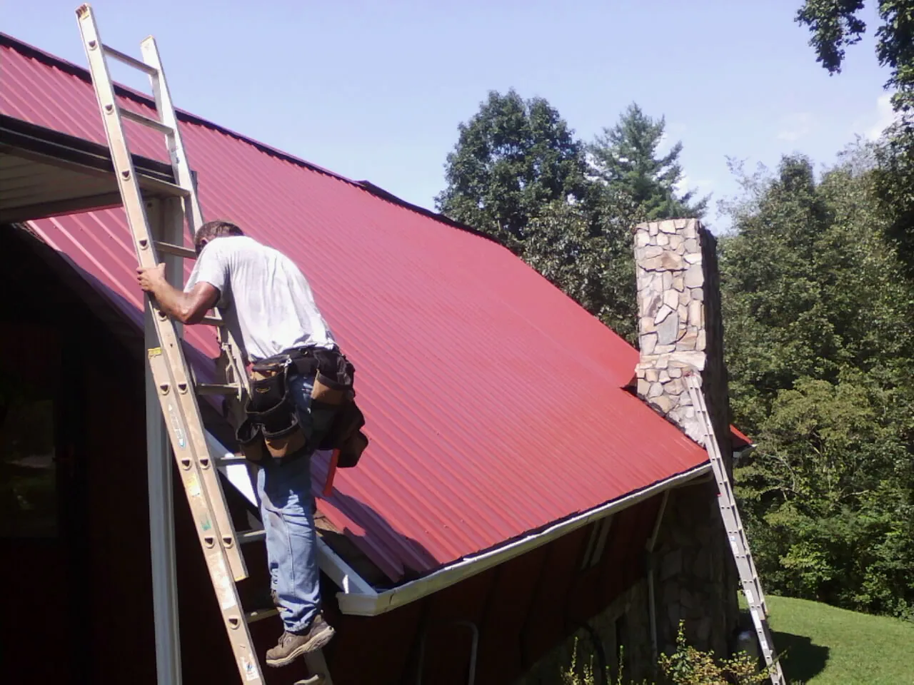 Roofer on a ladder beside a red metal roof and stone chimney — Soffit & Fascia Repair by Roofing Brothers in Cook Springs